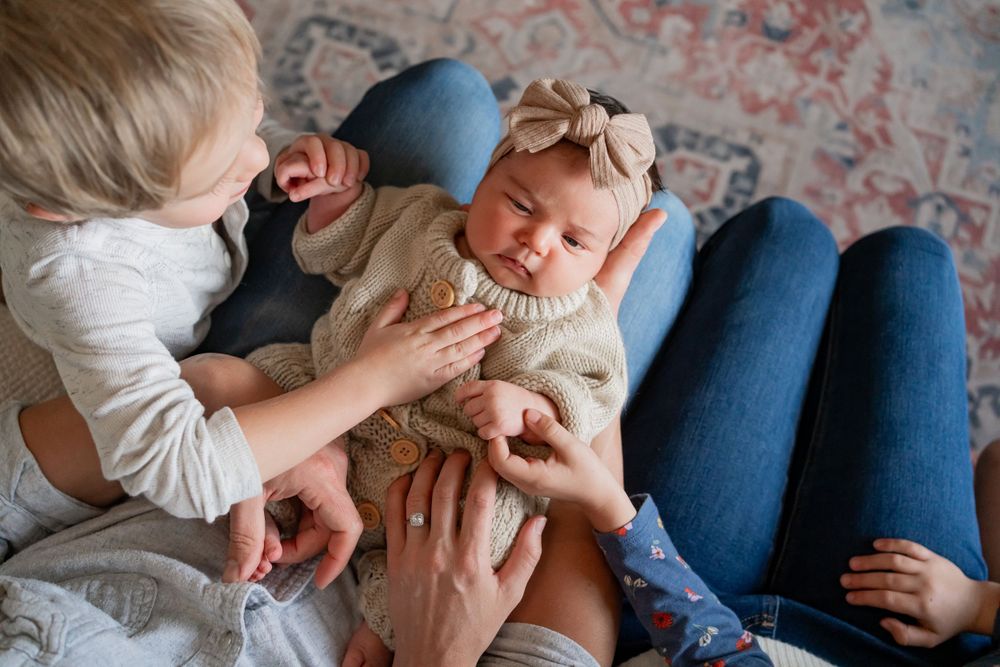 Siblings interact with a baby wearing a cream sweater and bow on a couch.