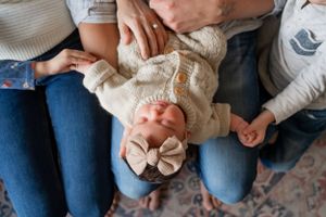 An infant wearing a cream sweater and bow lies across laps on a patterned rug.