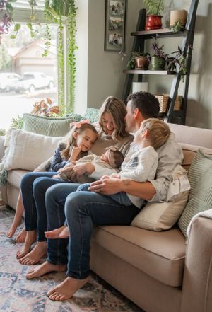 A family relaxes together on a couch near a window with hanging plants.