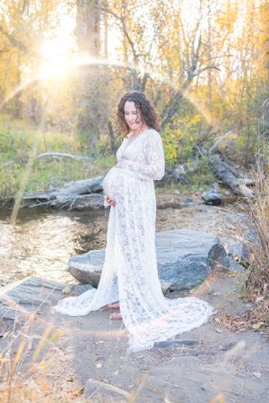 Maternity photo featuring woman in white lace gown by rocky stream.