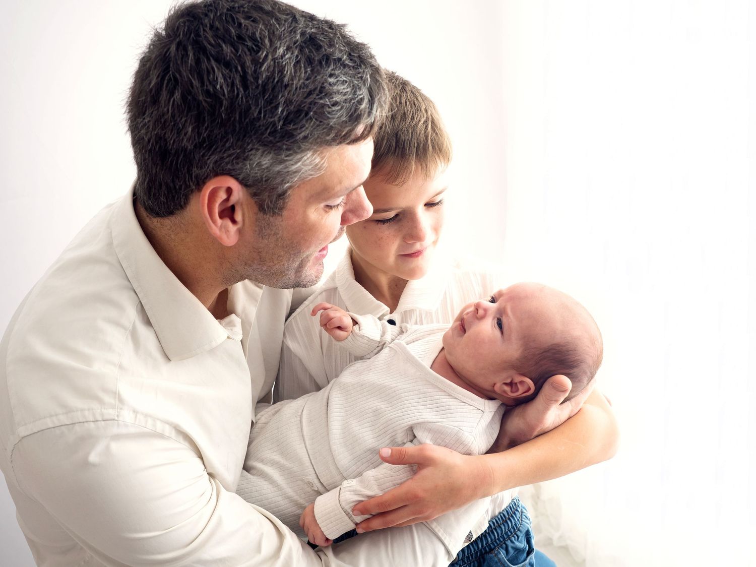 Photo of a father and young son holding a newborn baby and smiling. The light is coming through the white curtain behind