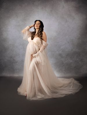 Elegant maternity portrait in flowing white gown against textured grey background.