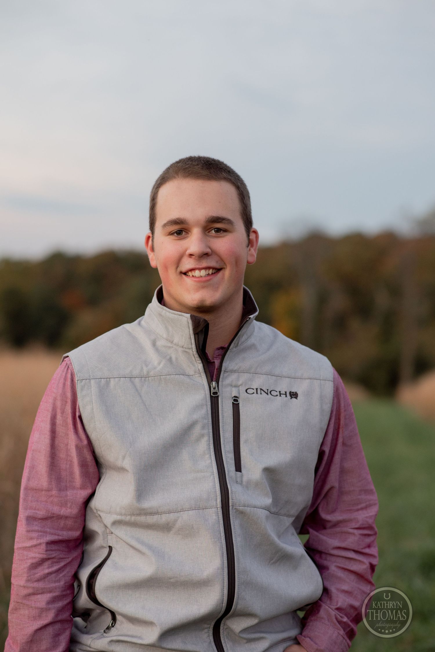 Person wearing gray vest and pink shirt smiles warmly against autumn outdoor background.