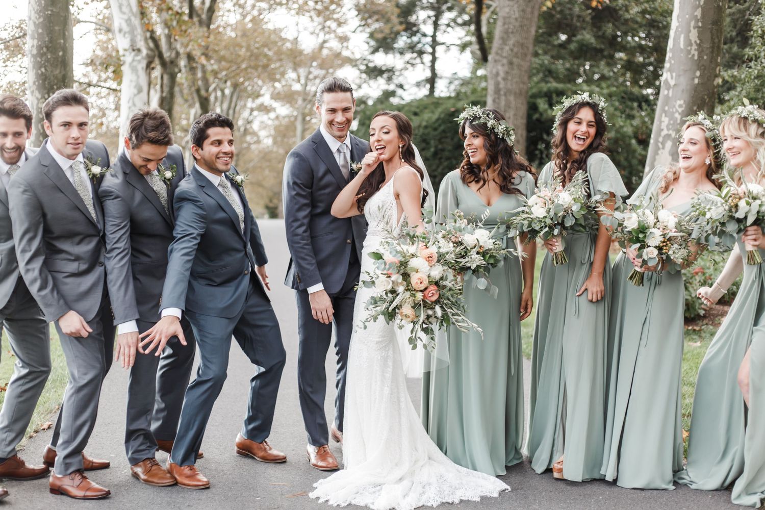 Wedding party poses playfully outdoors with bridesmaids in sage green dresses.
