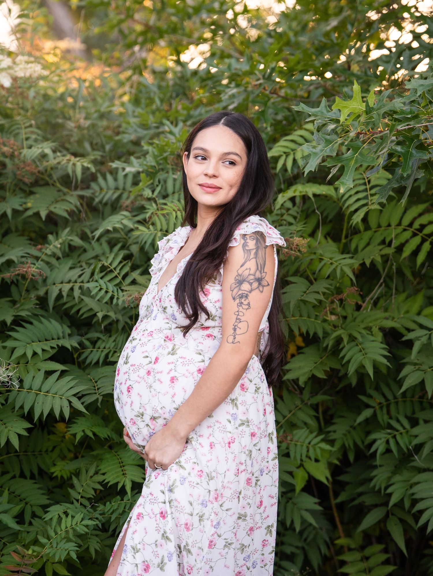 A pregnant woman in a floral dress poses in front of lush green foliage during a sunny outdoor photoshoot.