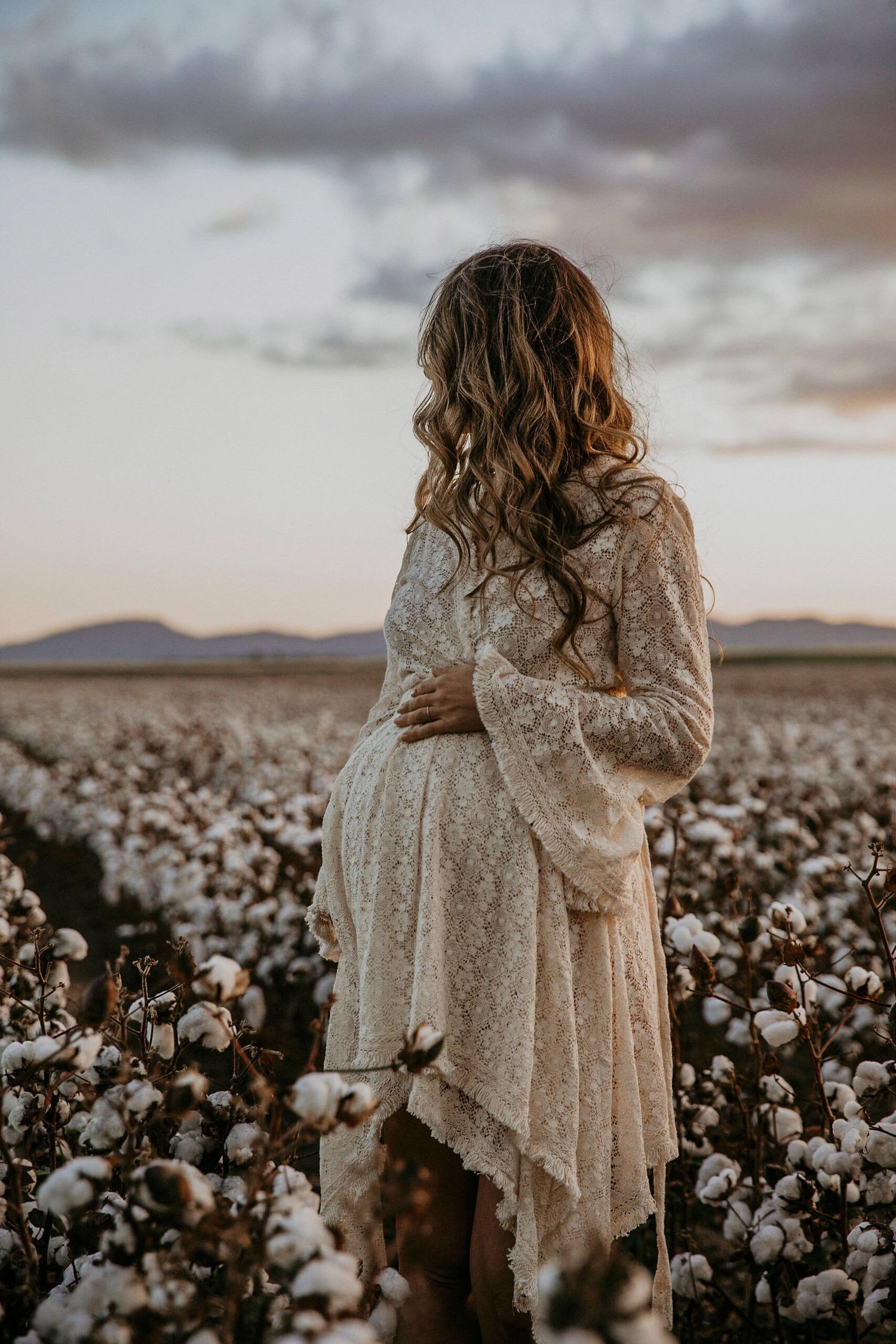 Pregnant woman in lace dress standing in cotton field at sunset.