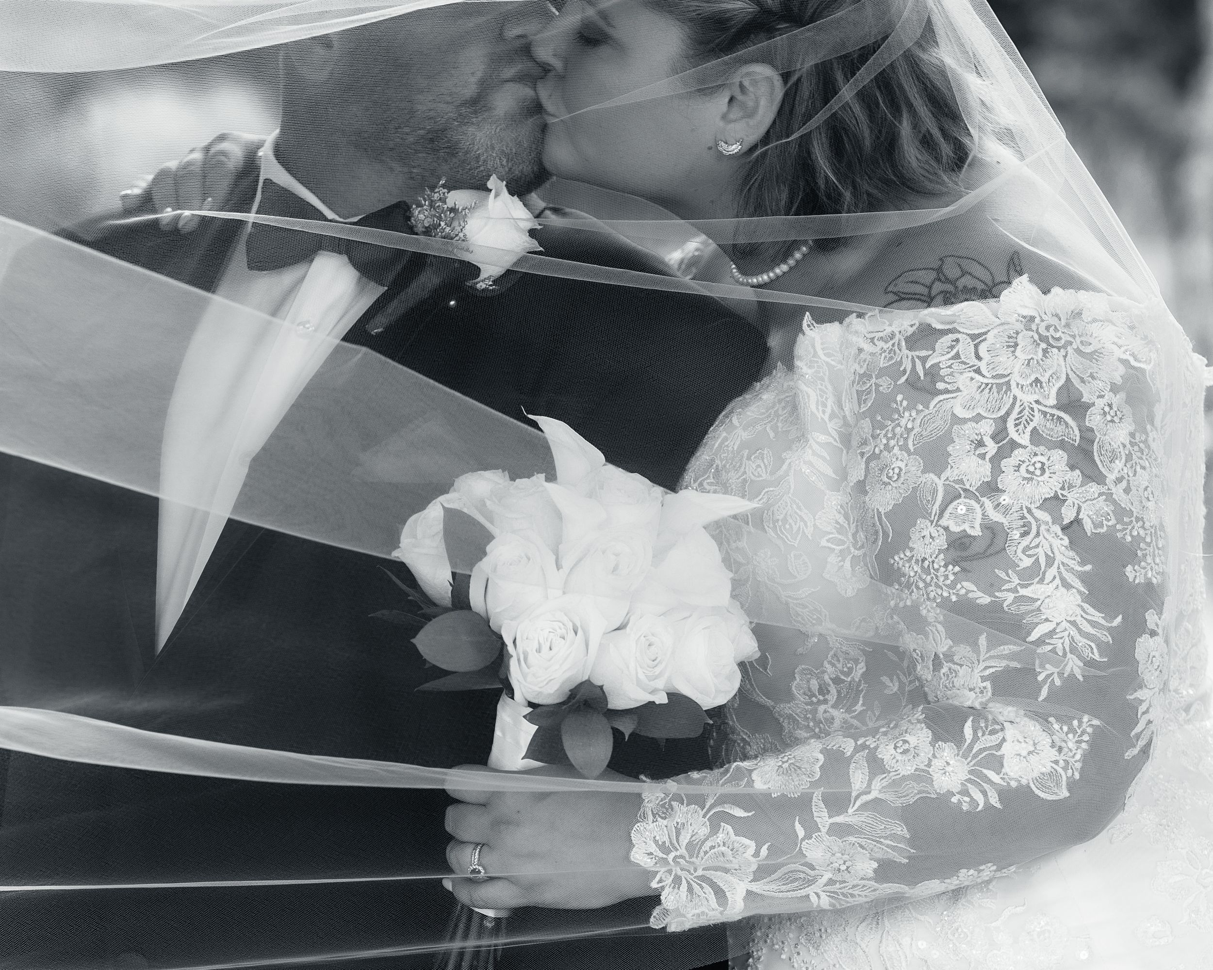 Black and white close-up of a wedding couple kissing under a flowing veil with lace details.