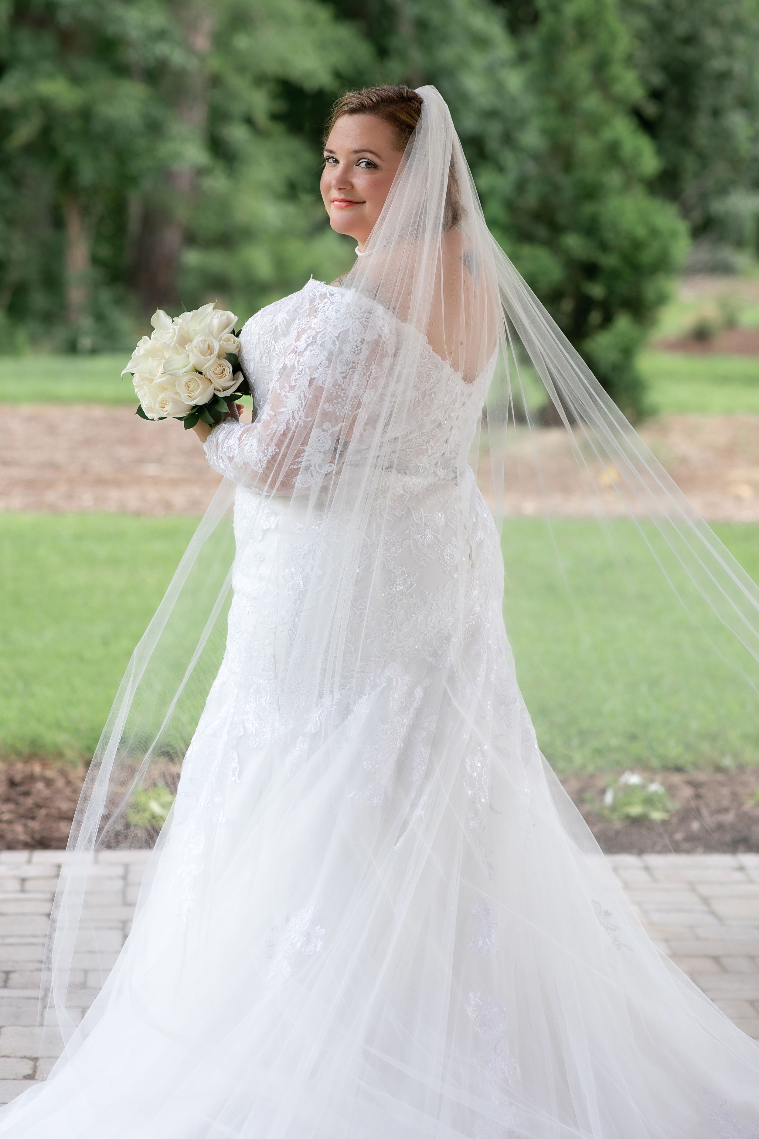 A bride in a white lace wedding dress poses with her bouquet in an outdoor garden setting.