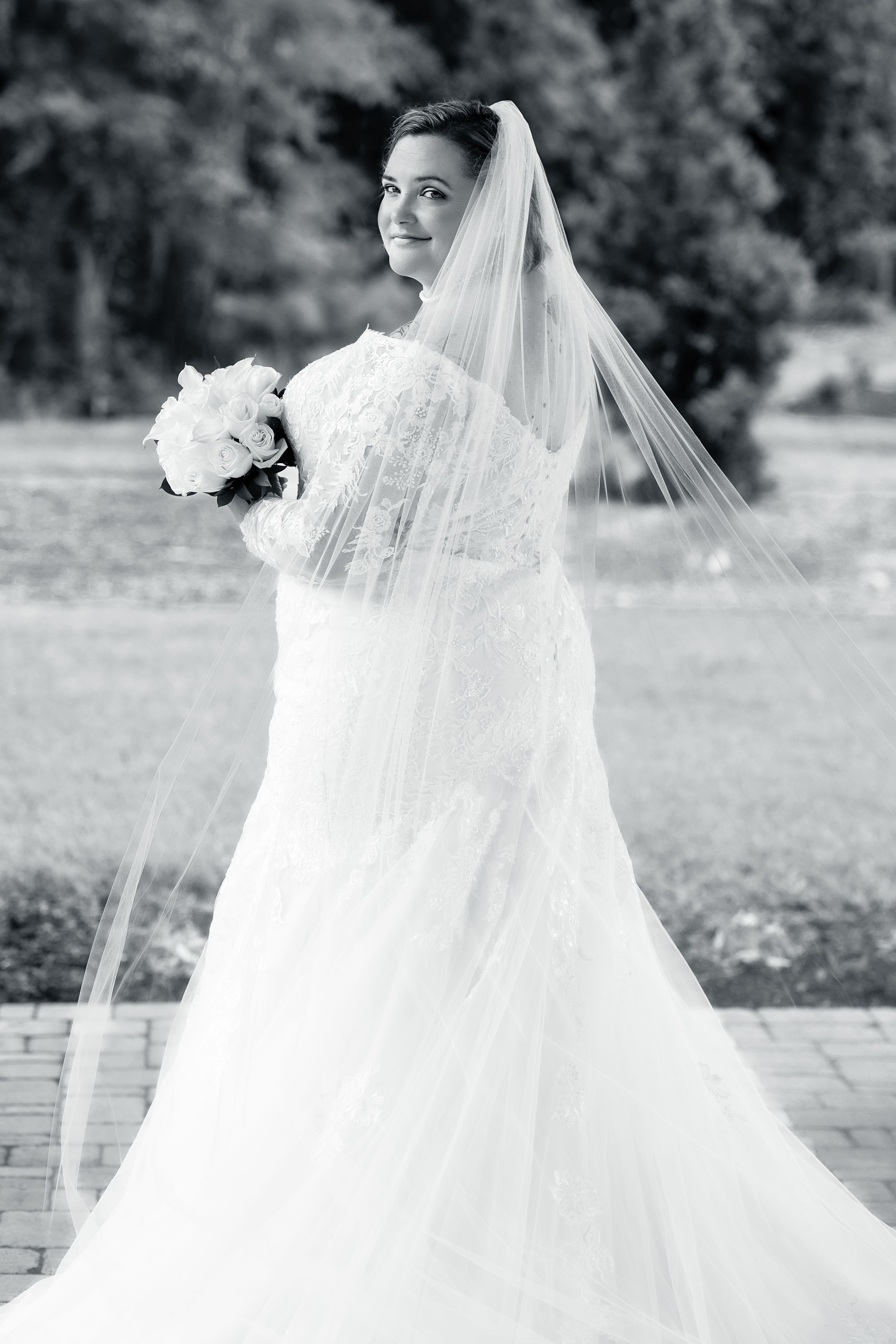Black and white portrait of a bride in a lace wedding gown with flowing veil holding a white bouquet.