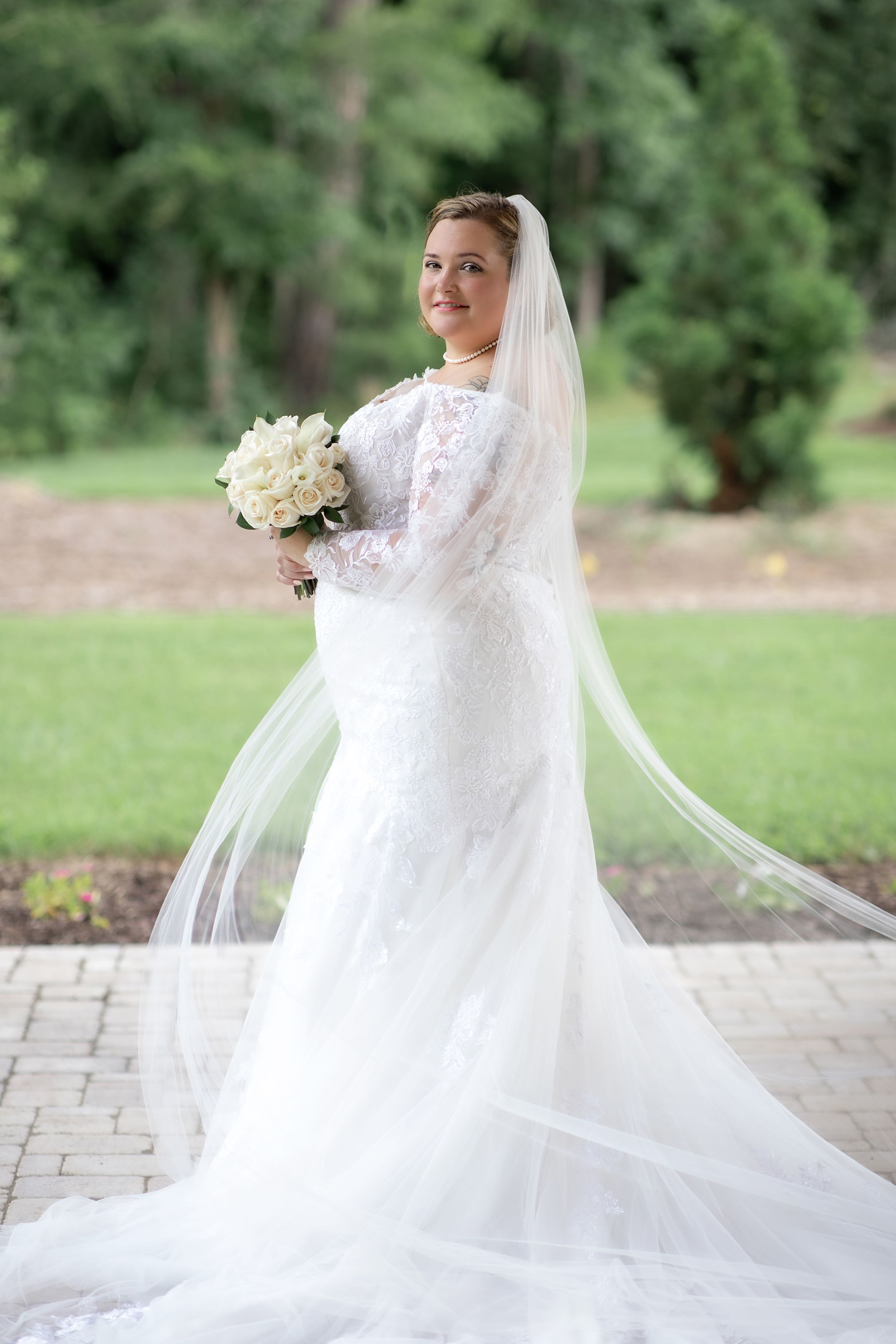 A radiant bride poses in her full-length lace wedding gown with cathedral veil on a stone path.
