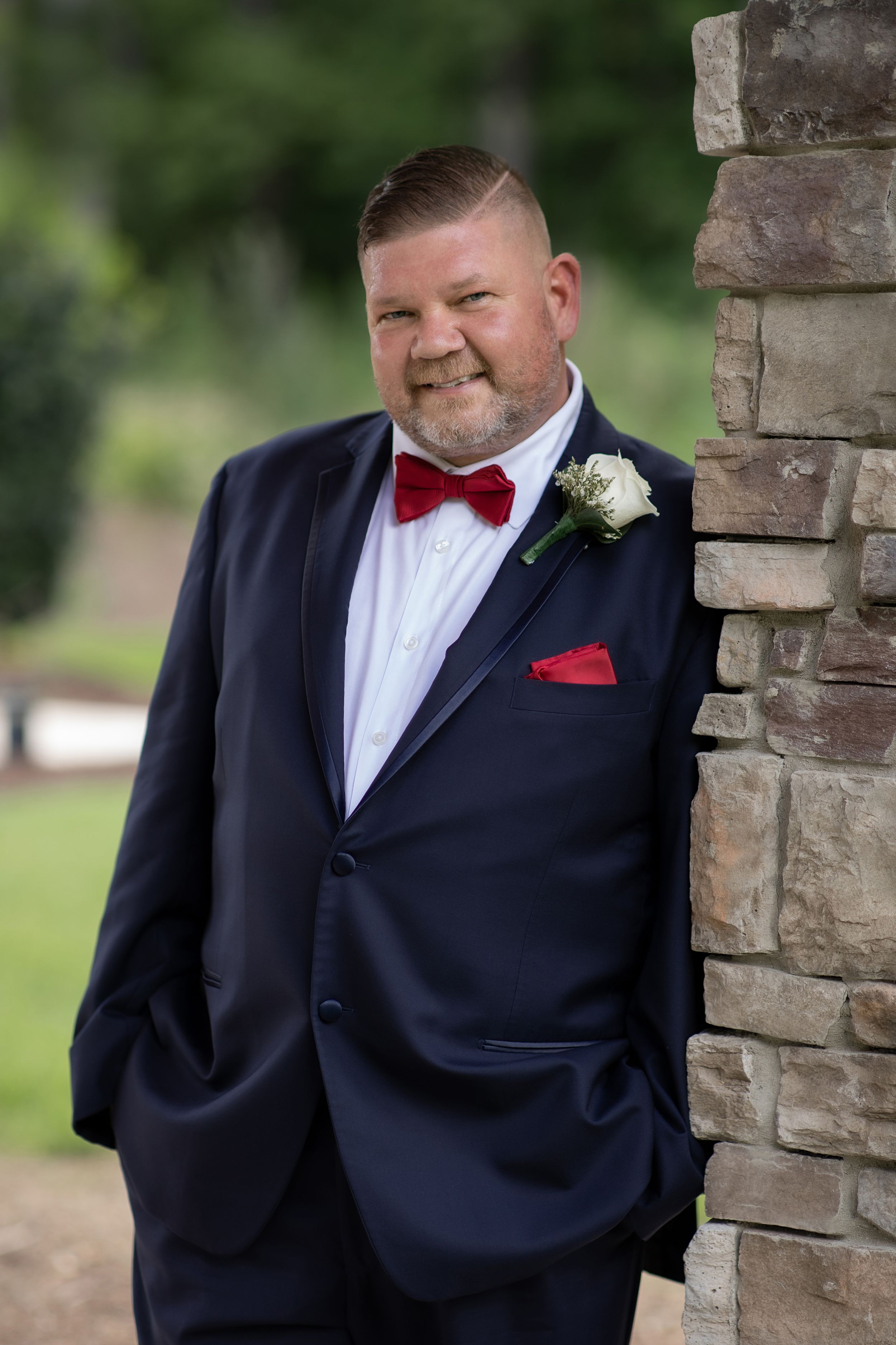 Portrait of a groom in a navy suit and red bow tie leaning against a stone pillar.
