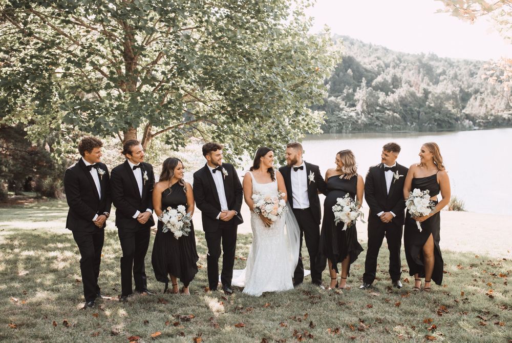 Wedding party in formal attire posed together on grassy hillside near lake.