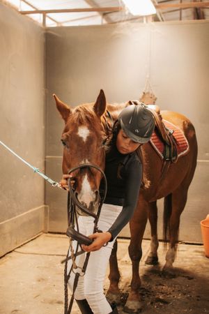 Series showing person in riding gear tending to saddled brown horse in indoor arena.