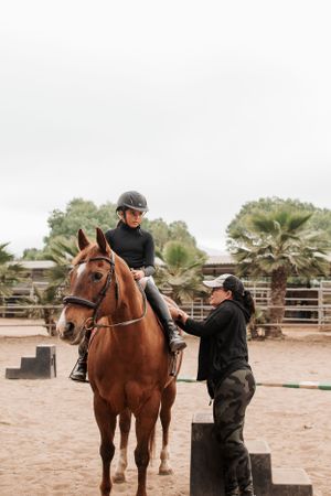 Horse with riding equipment stands in sandy arena while being assisted.