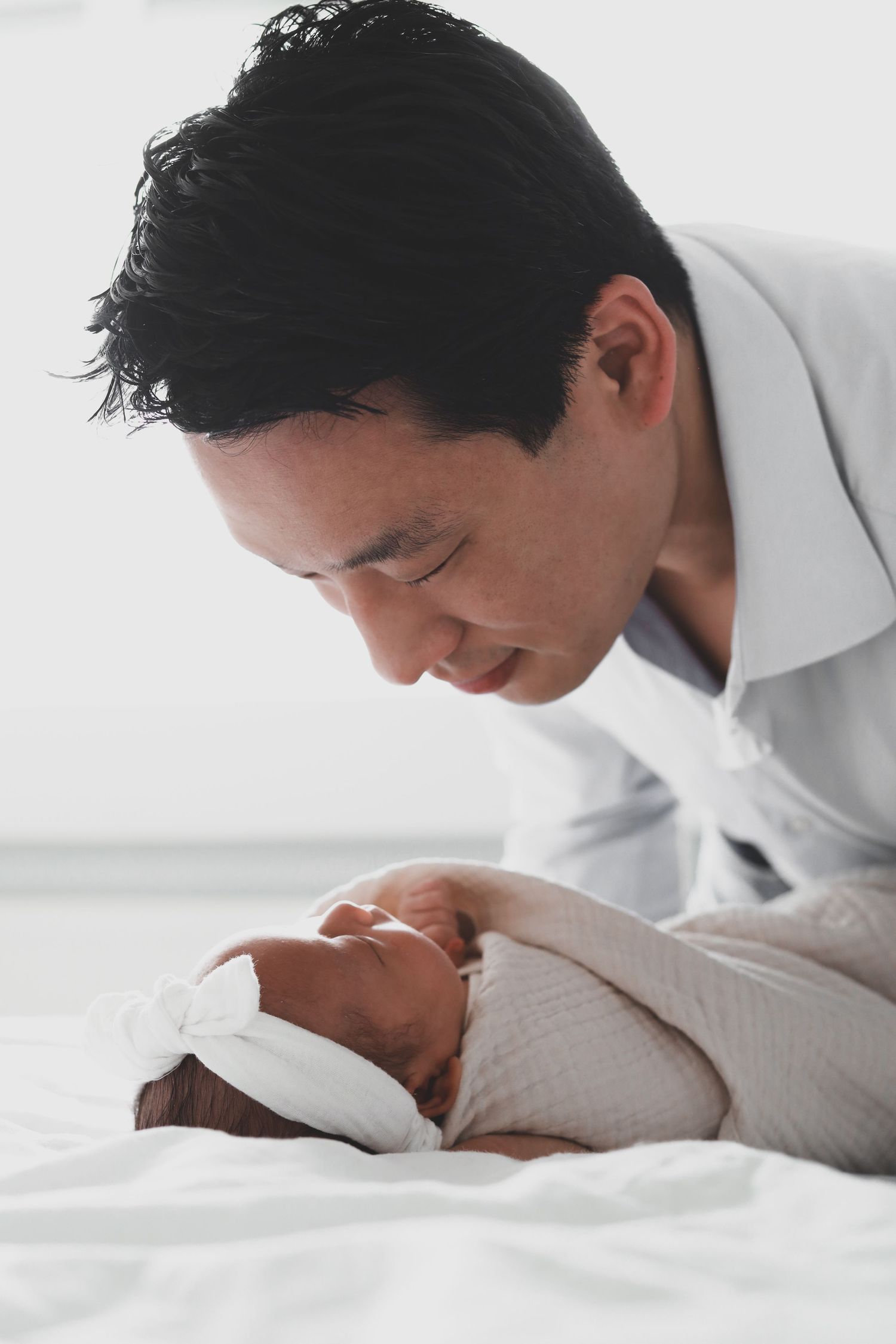 Asian father leaning over smiling newborn baby lying on white bed.