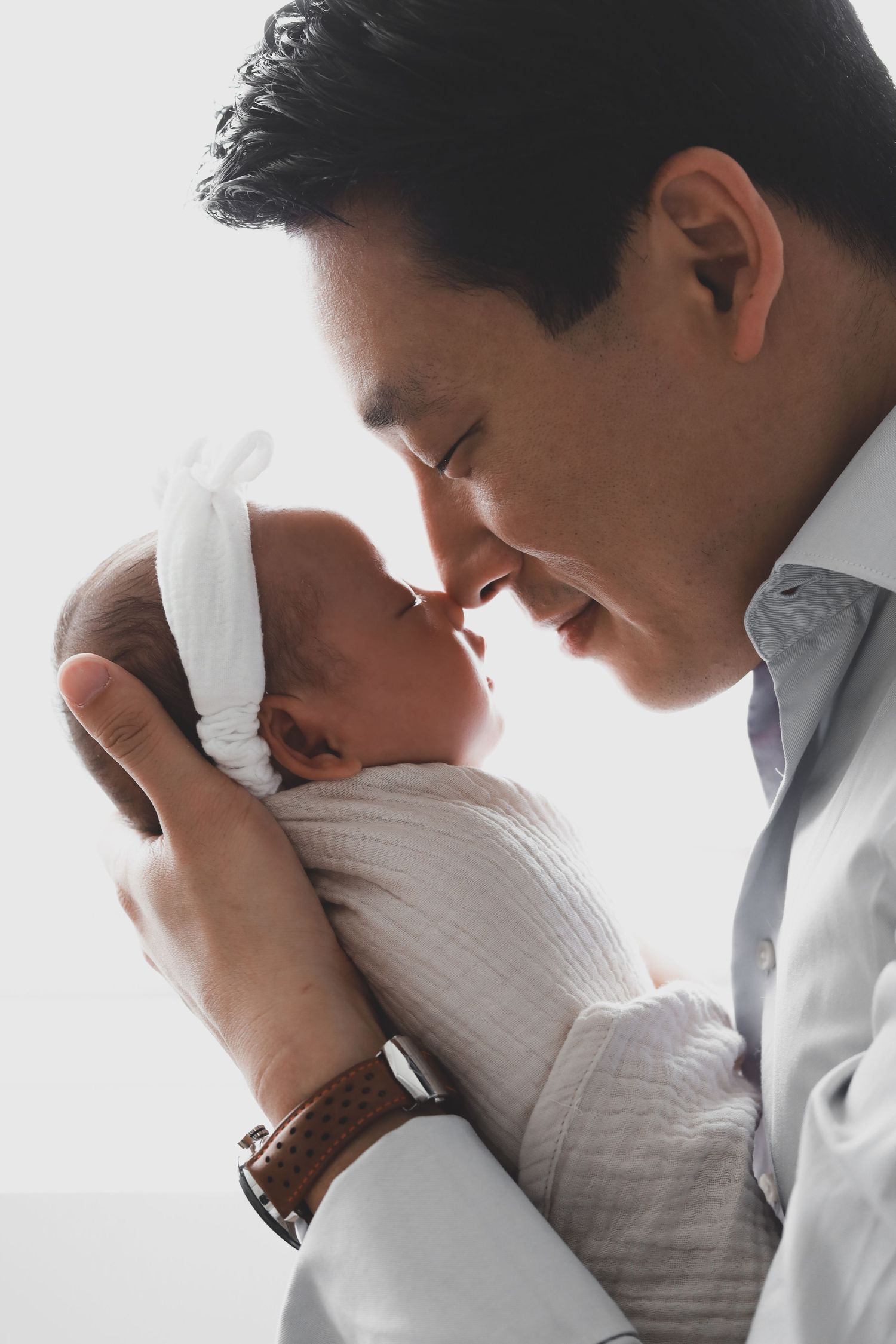 Father touching noses with swaddled newborn baby in white headband.