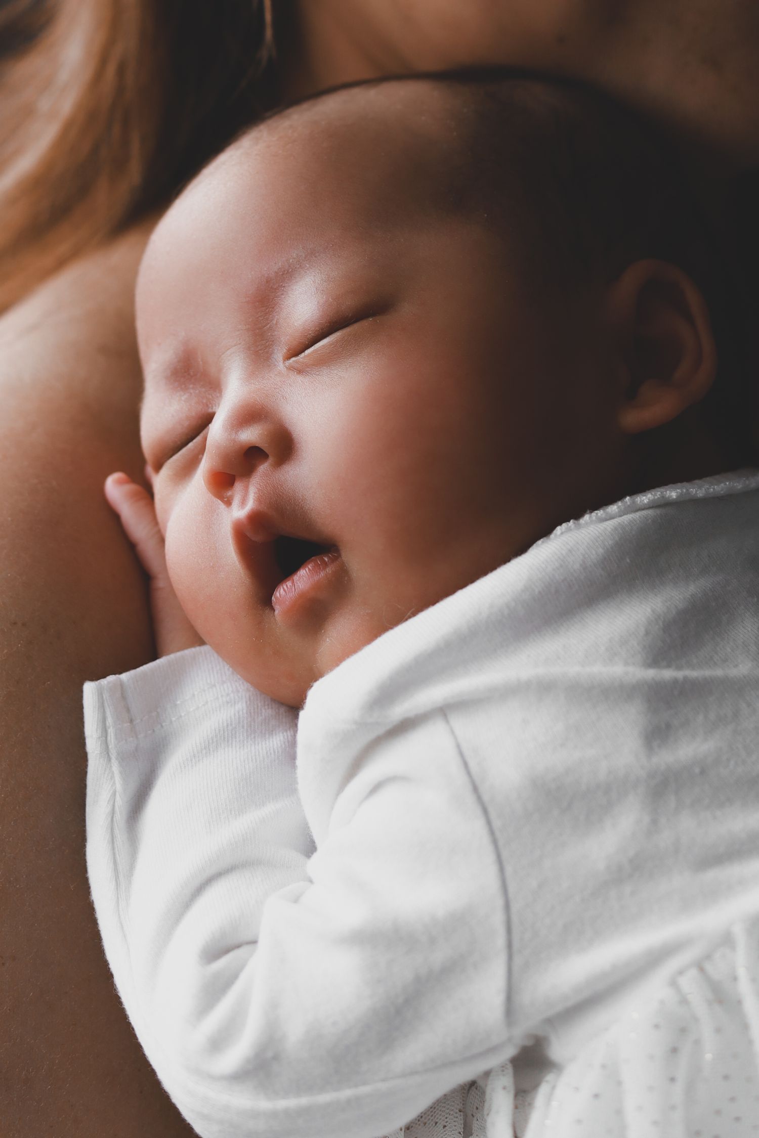 Peaceful Asian newborn baby sleeping with mouth open in white onesie.