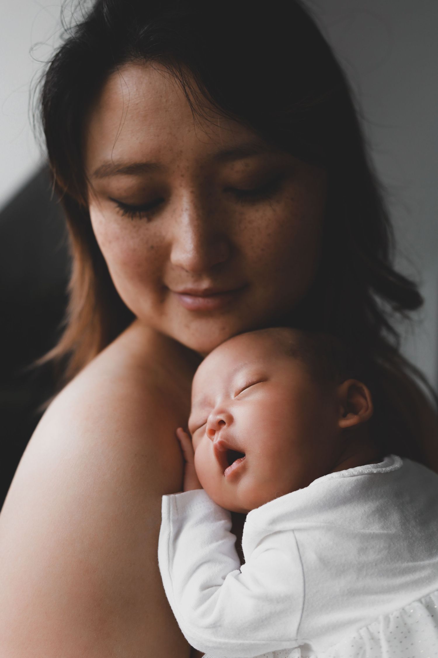 Asian mother smiling softly while holding her sleeping newborn baby close.