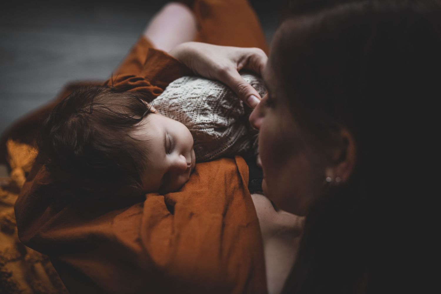Mother in rust orange dress gazing lovingly at sleeping newborn baby.