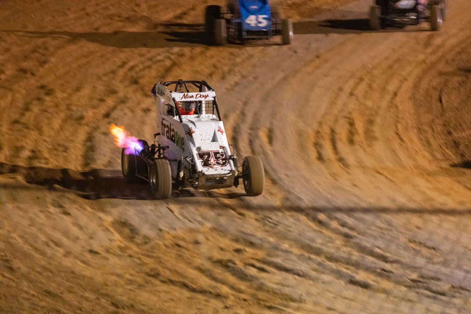 Sprint car racing on a dirt track at night with visible exhaust flames.