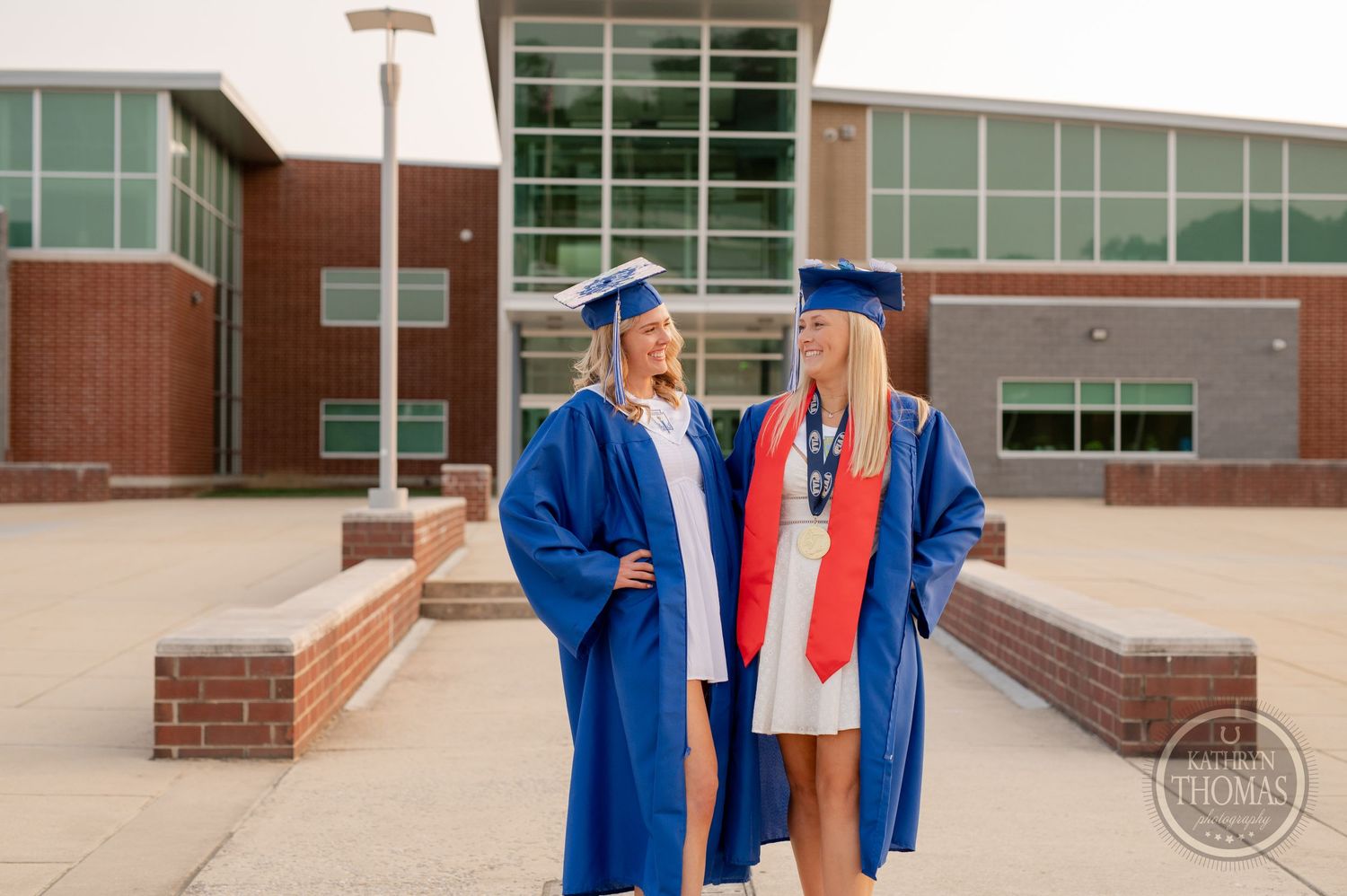 Two graduates in blue caps and gowns pose together outside brick school building entrance.