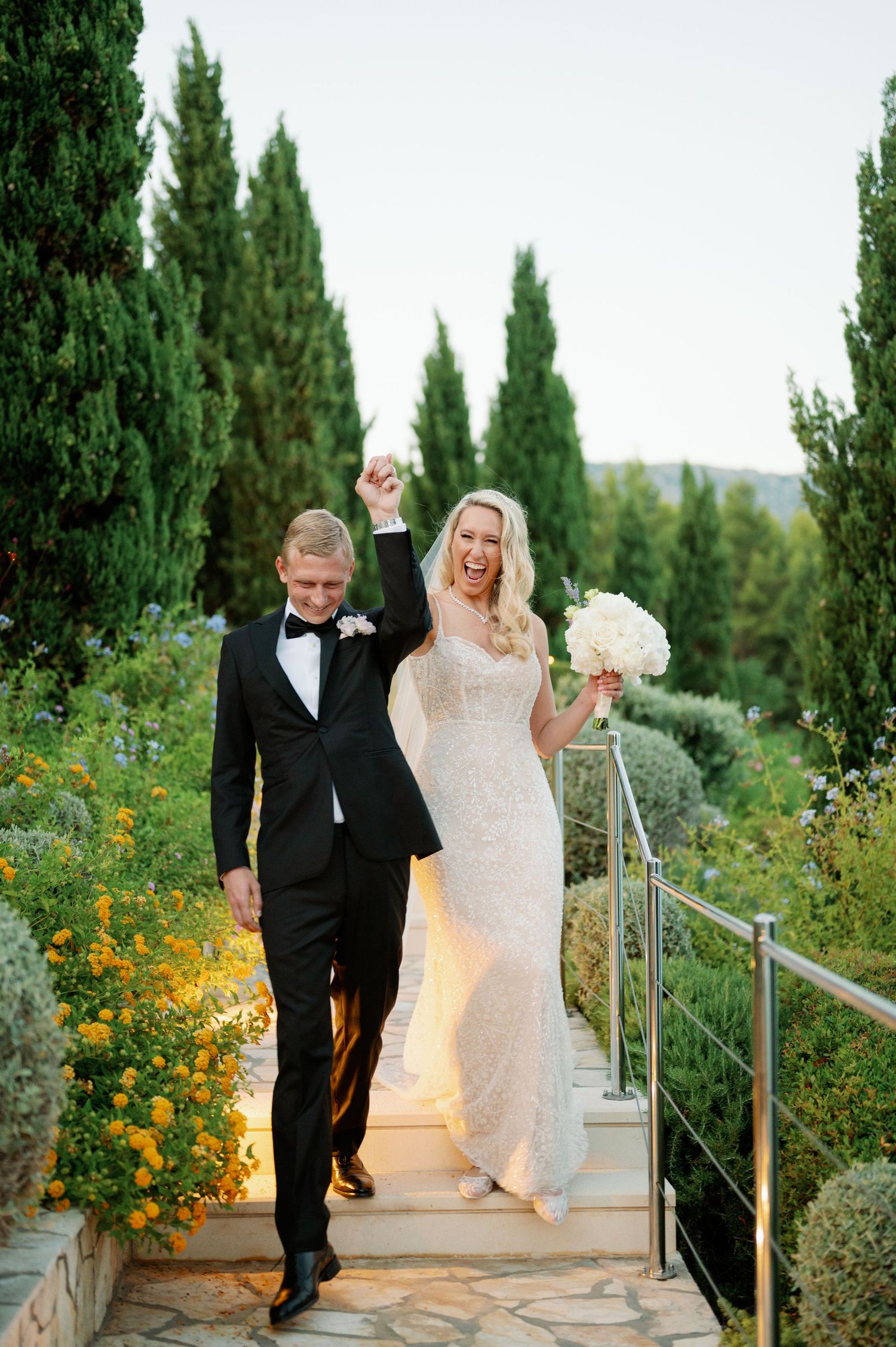 A couple celebrates on stone steps surrounded by cypress trees and yellow wildflowers.