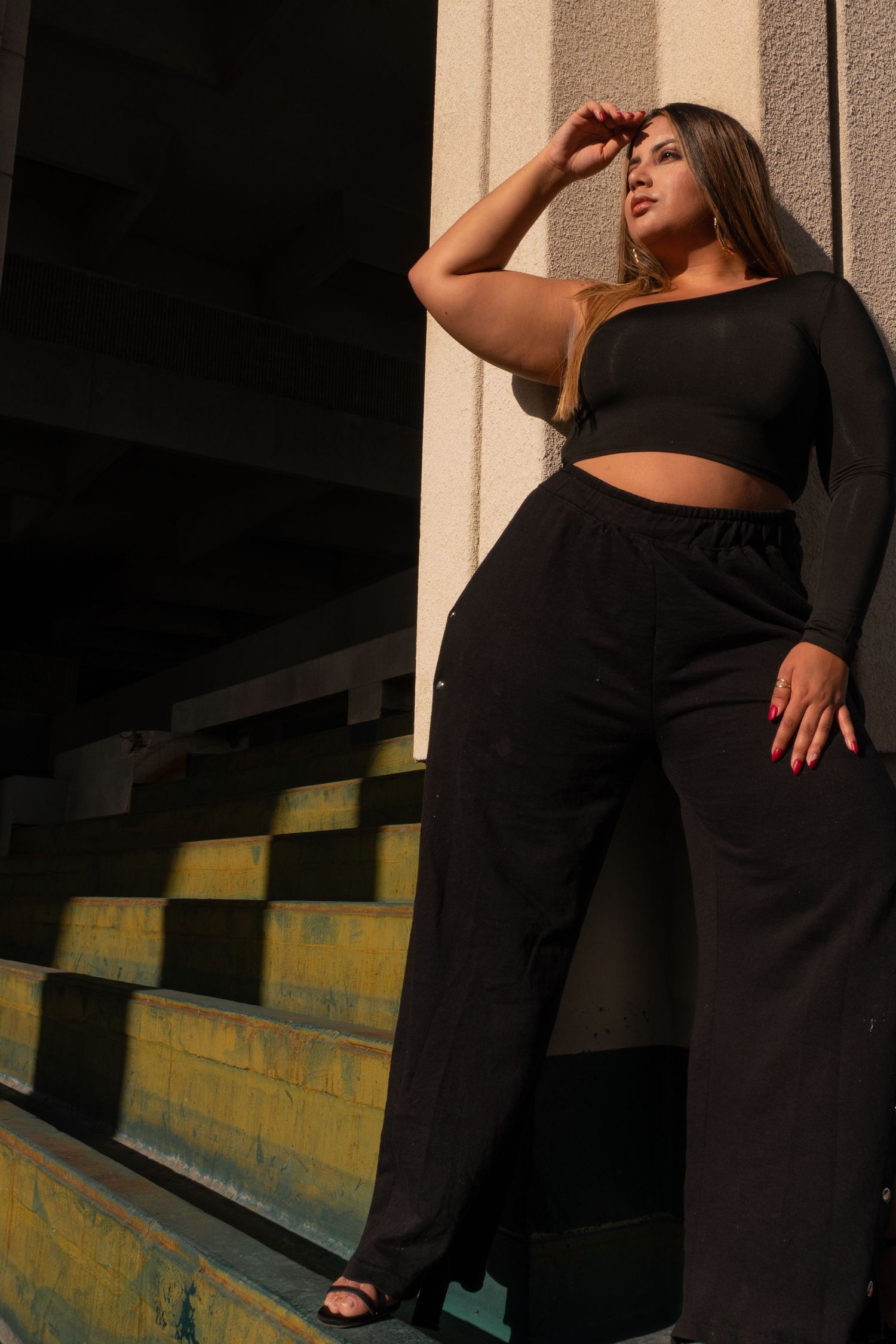 A plus-size woman poses confidently in a black crop top and wide-leg pants on outdoor steps in golden sunlight.