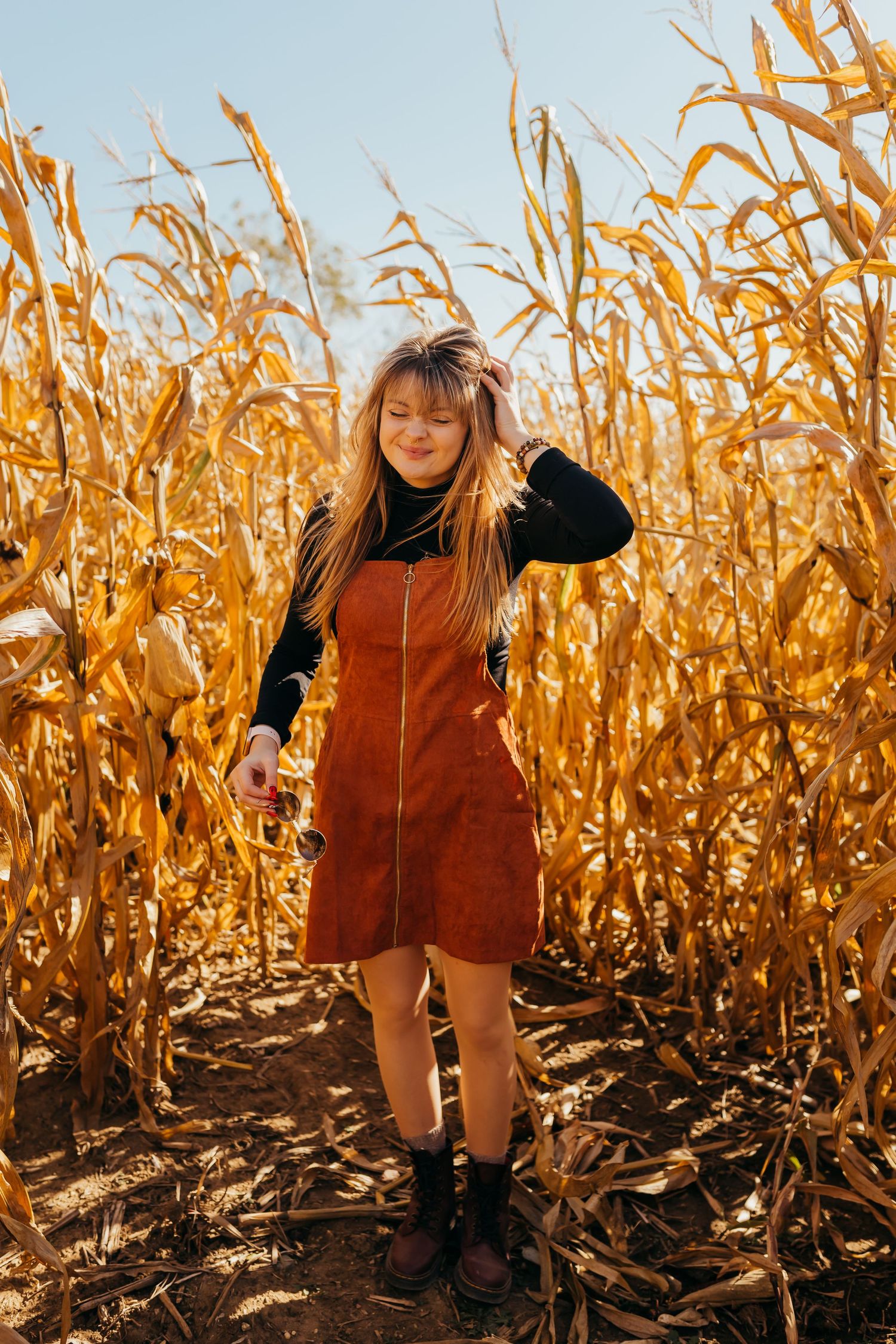 A person in a corduroy dress poses playfully in a sunny cornfield during autumn harvest season.