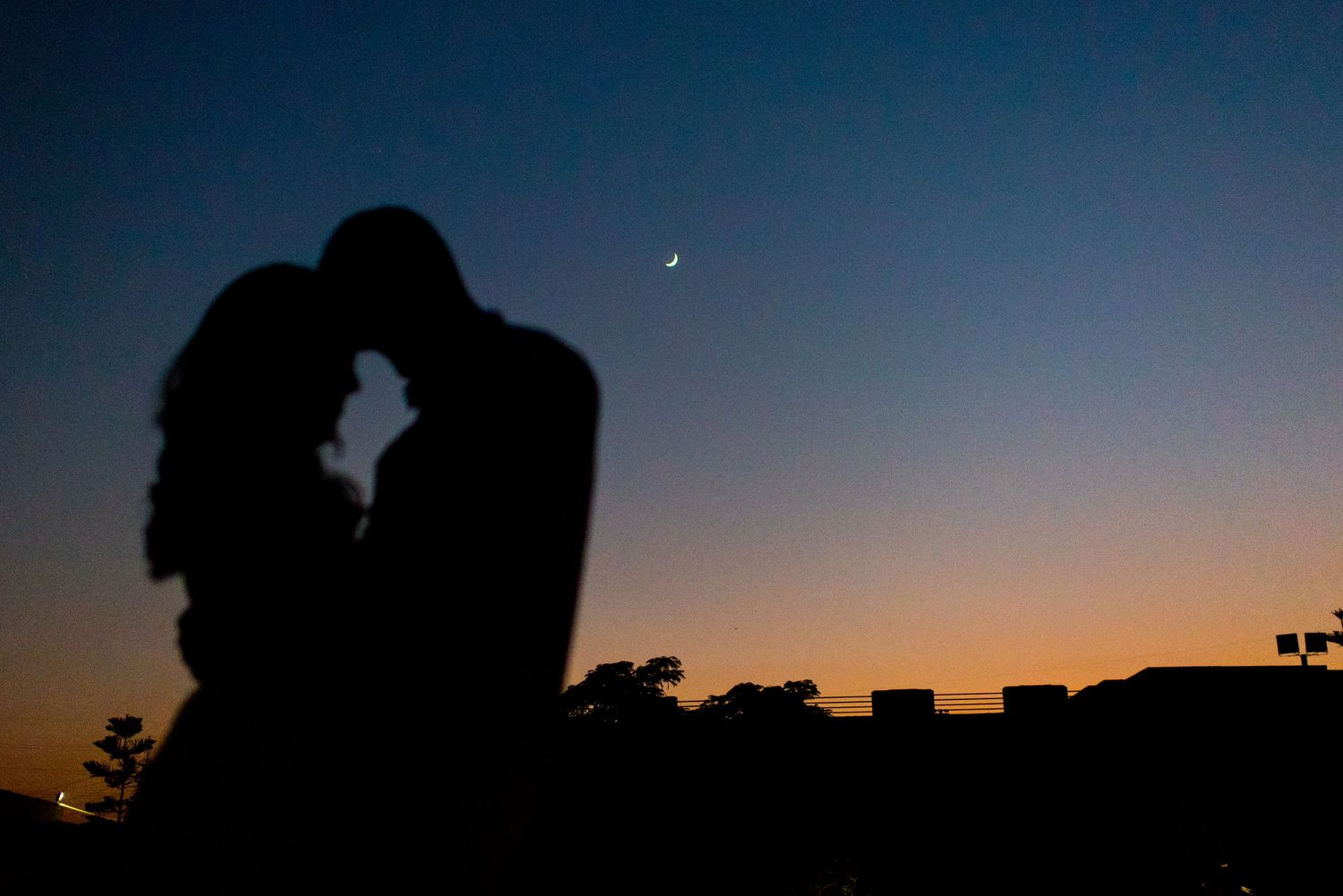 Romantic silhouette of a couple embracing against a twilight sky with a crescent moon visible in the background.