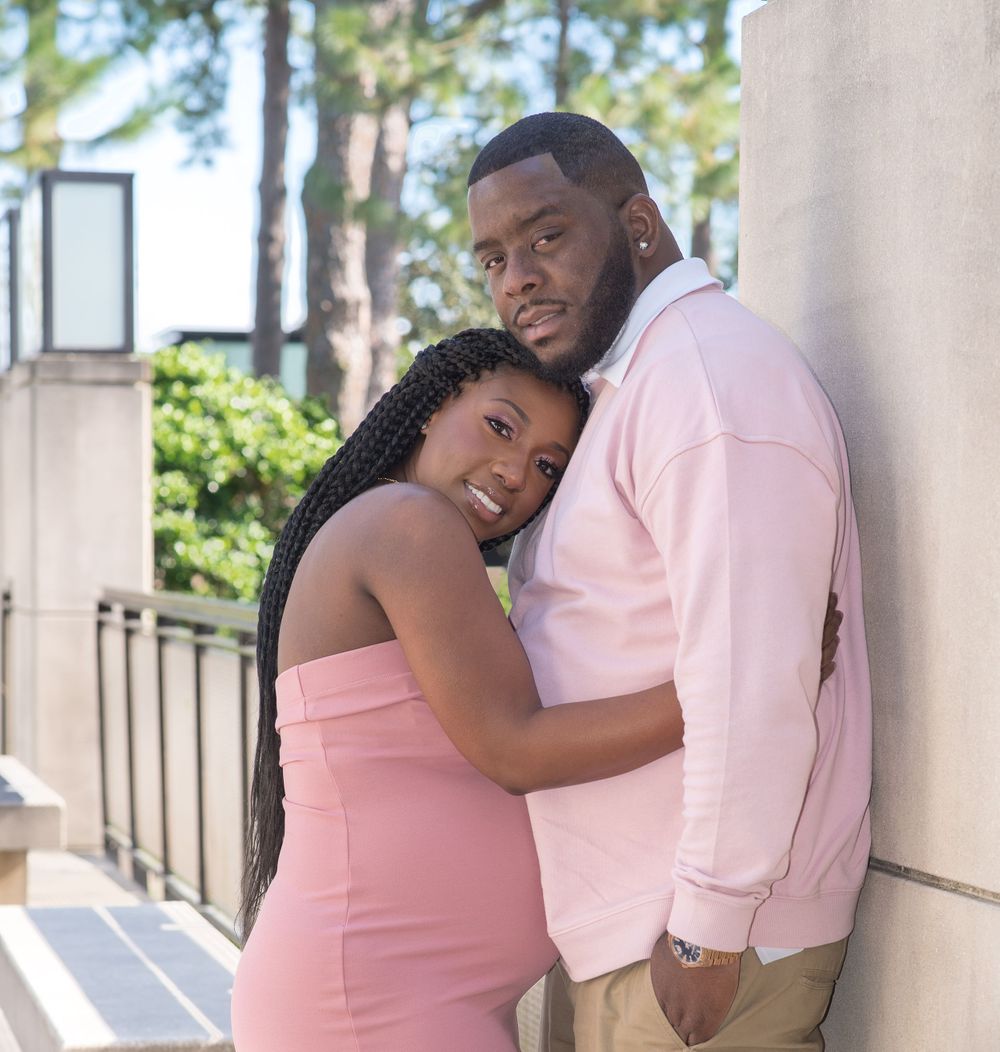 A couple wearing pink and white outfits pose affectionately against a building exterior.