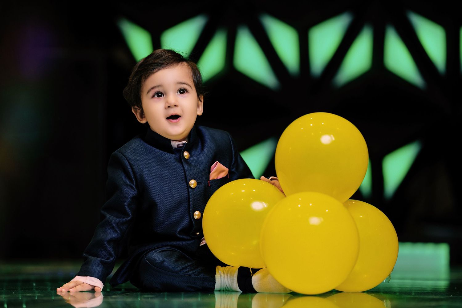 Young child in navy blue outfit poses with yellow balloons against geometric backdrop.