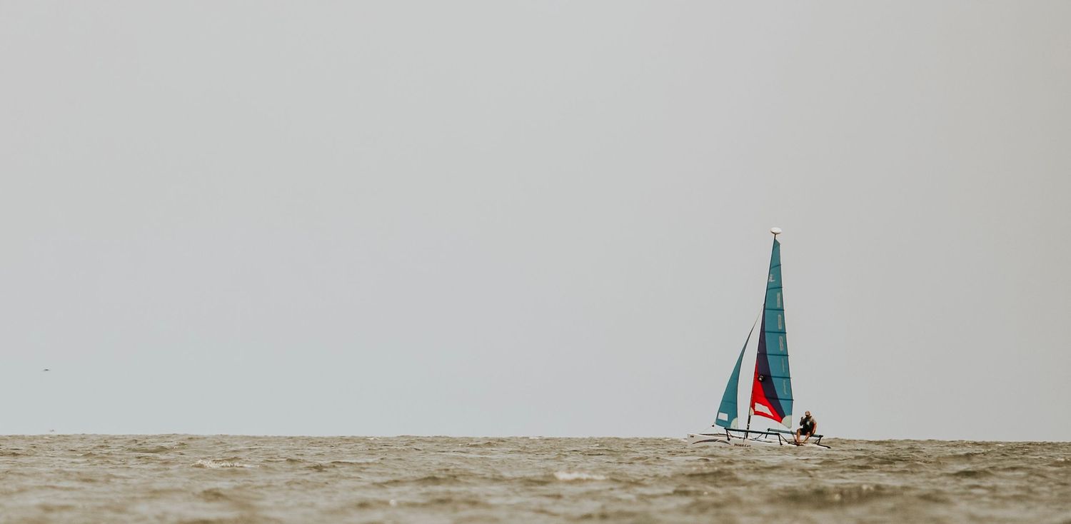 Sailboat with teal sails glides across misty waters against an overcast sky.