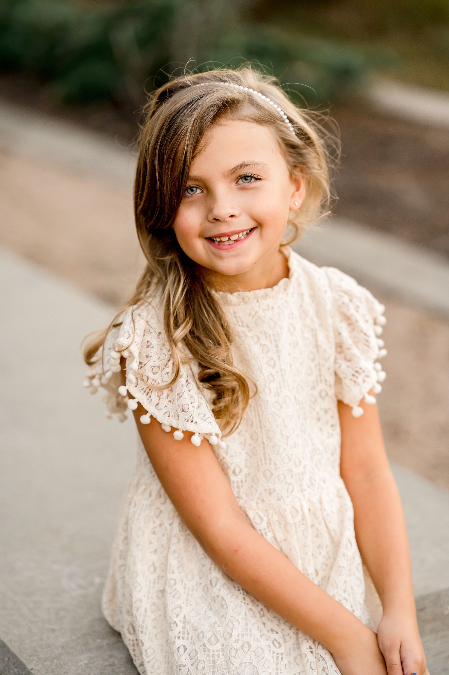 A child in a white lace dress smiles brightly in an outdoor portrait session.