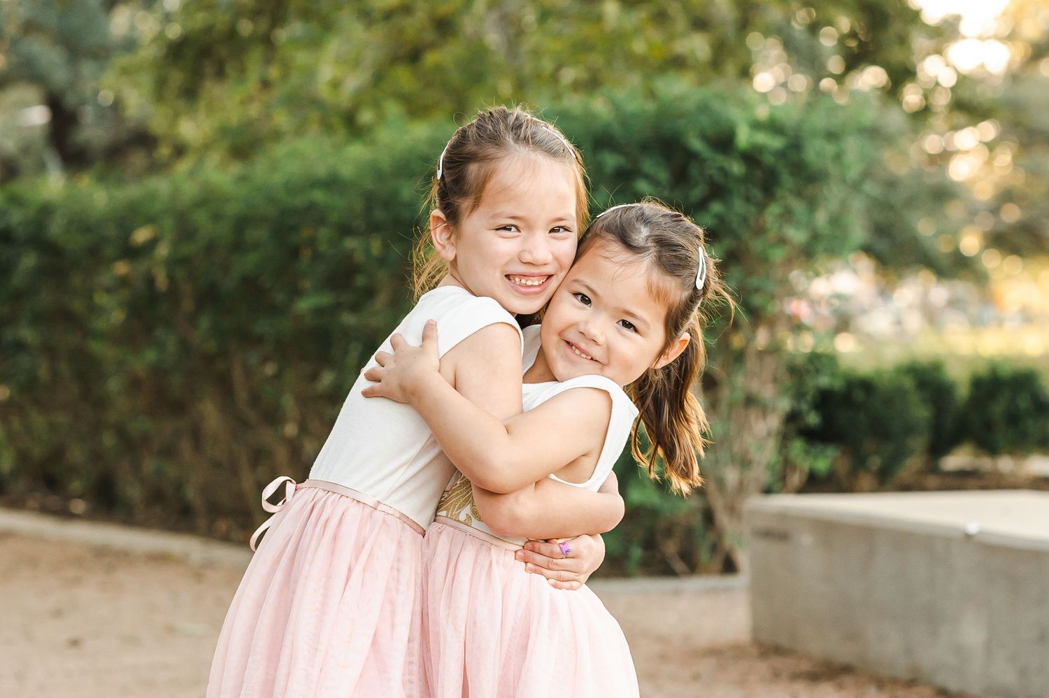 Two young girls in pink dresses share a sweet embrace while standing on a stone path outdoors.