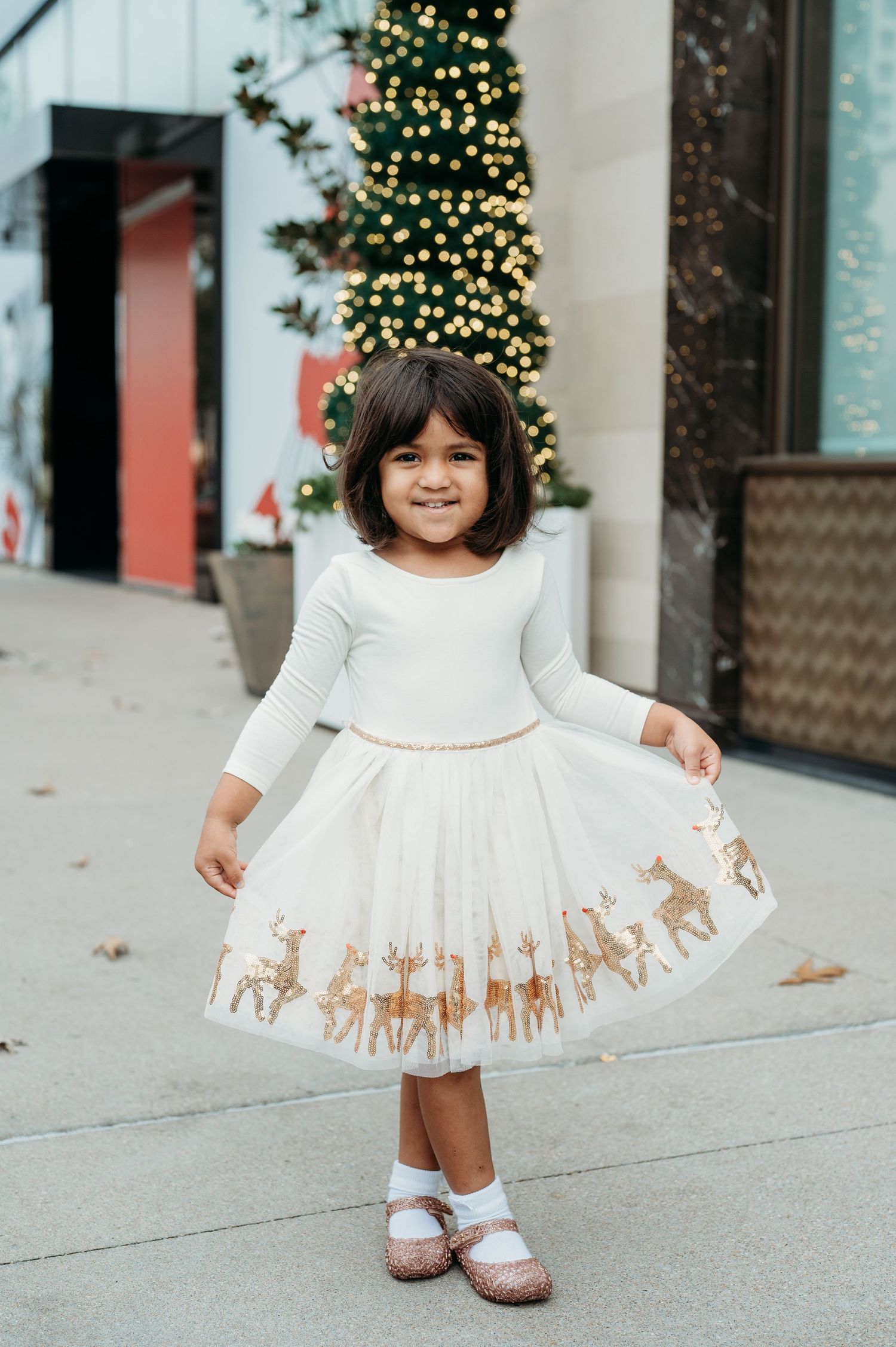 White dress with gold reindeer pattern twirls on a sidewalk near holiday decorations.
