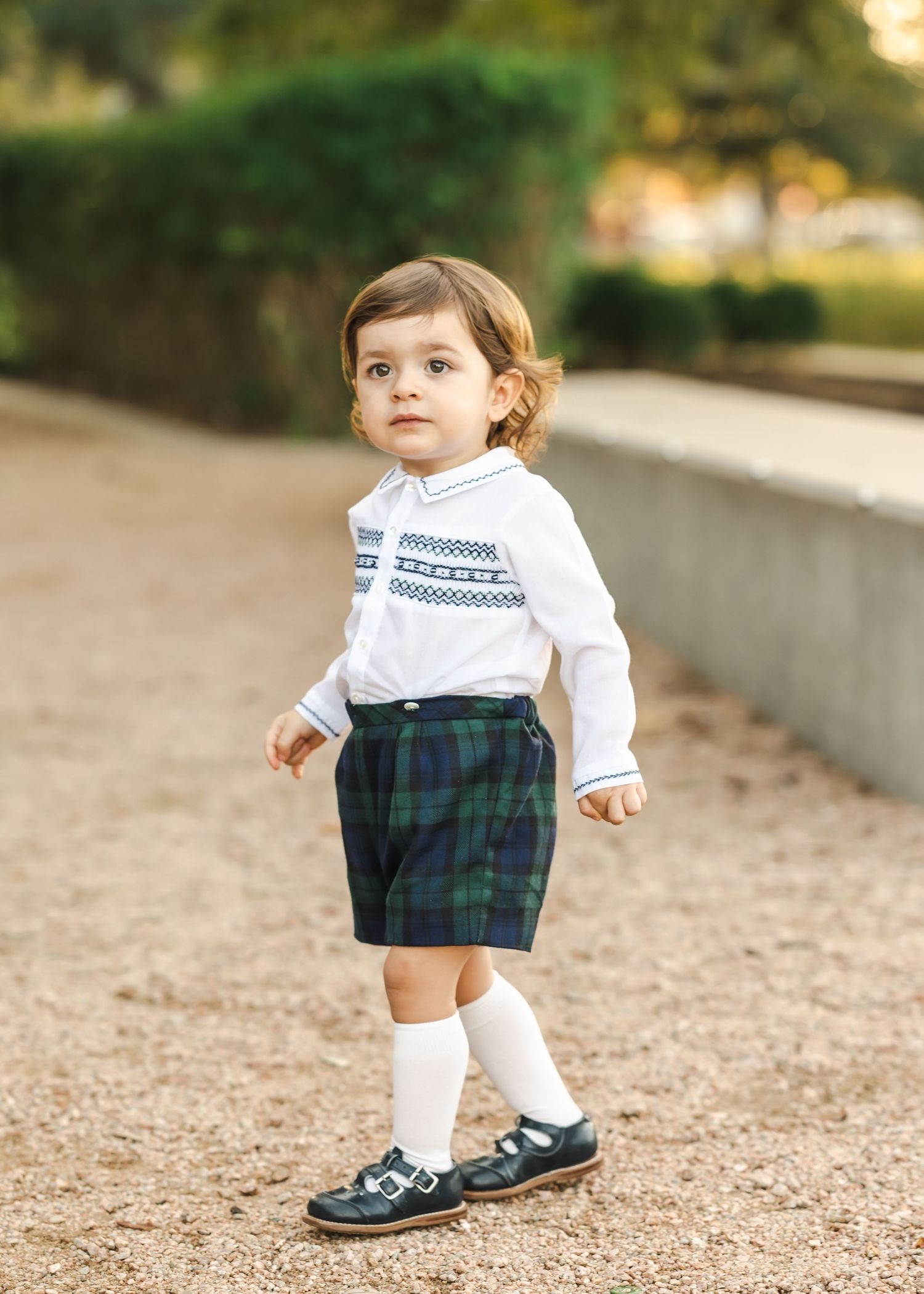 Small child in plaid outfit takes steps on gravel path during photo session.