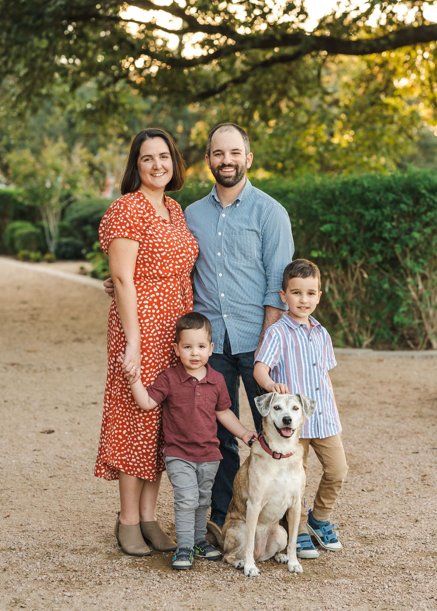 A family of four poses with their white dog on a dirt path at sunset with green foliage in background.