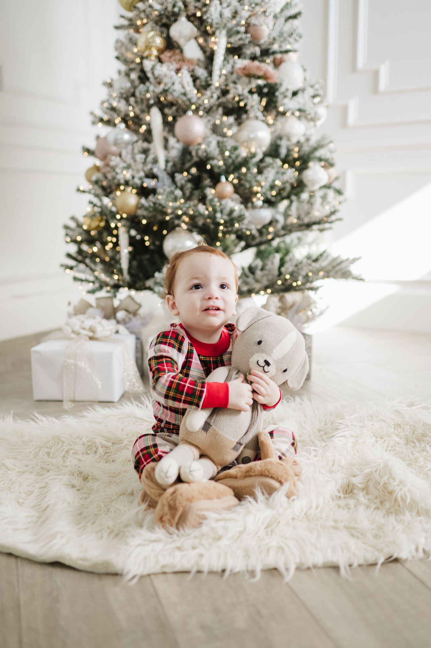A toddler in plaid pajamas hugs a stuffed animal in front of a decorated Christmas tree with wrapped presents.