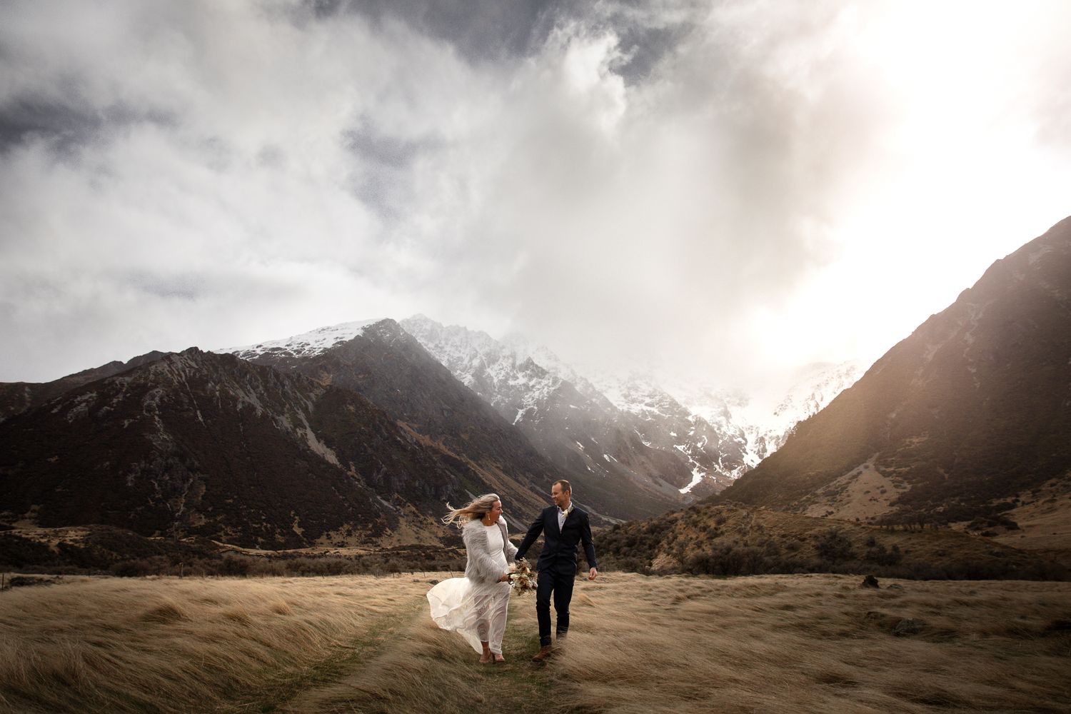 Couple walking hand in hand through dramatic mountain valley with snow-capped peaks.