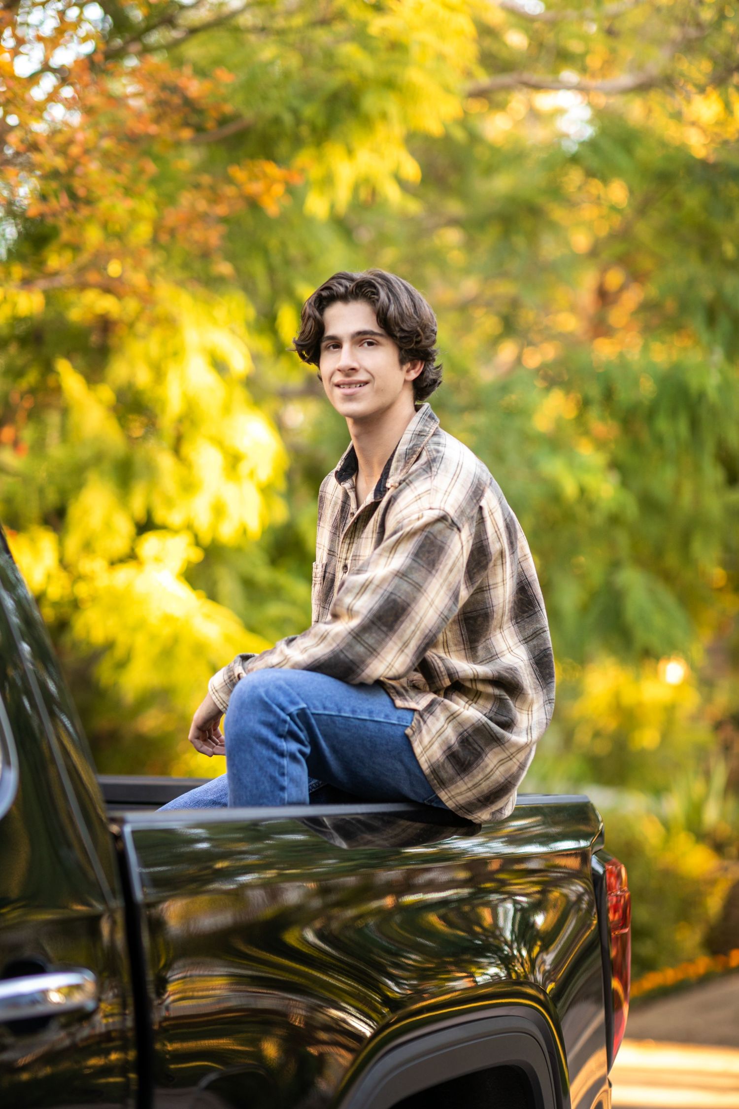 A high school senior in plaid shirt sitting on black truck bed with autumn foliage background.