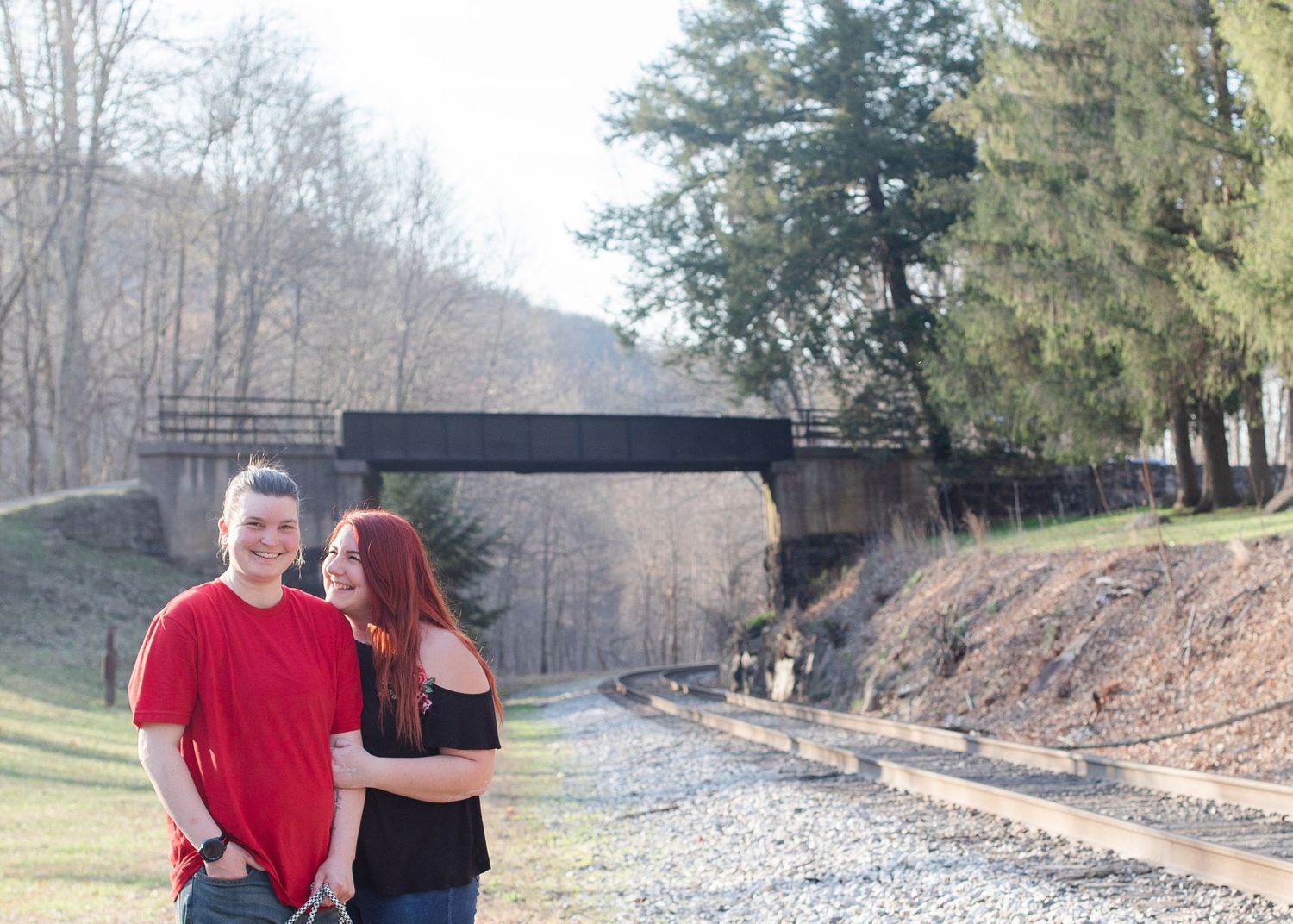 A couple poses near a rustic bridge and walking path surrounded by winter trees.
