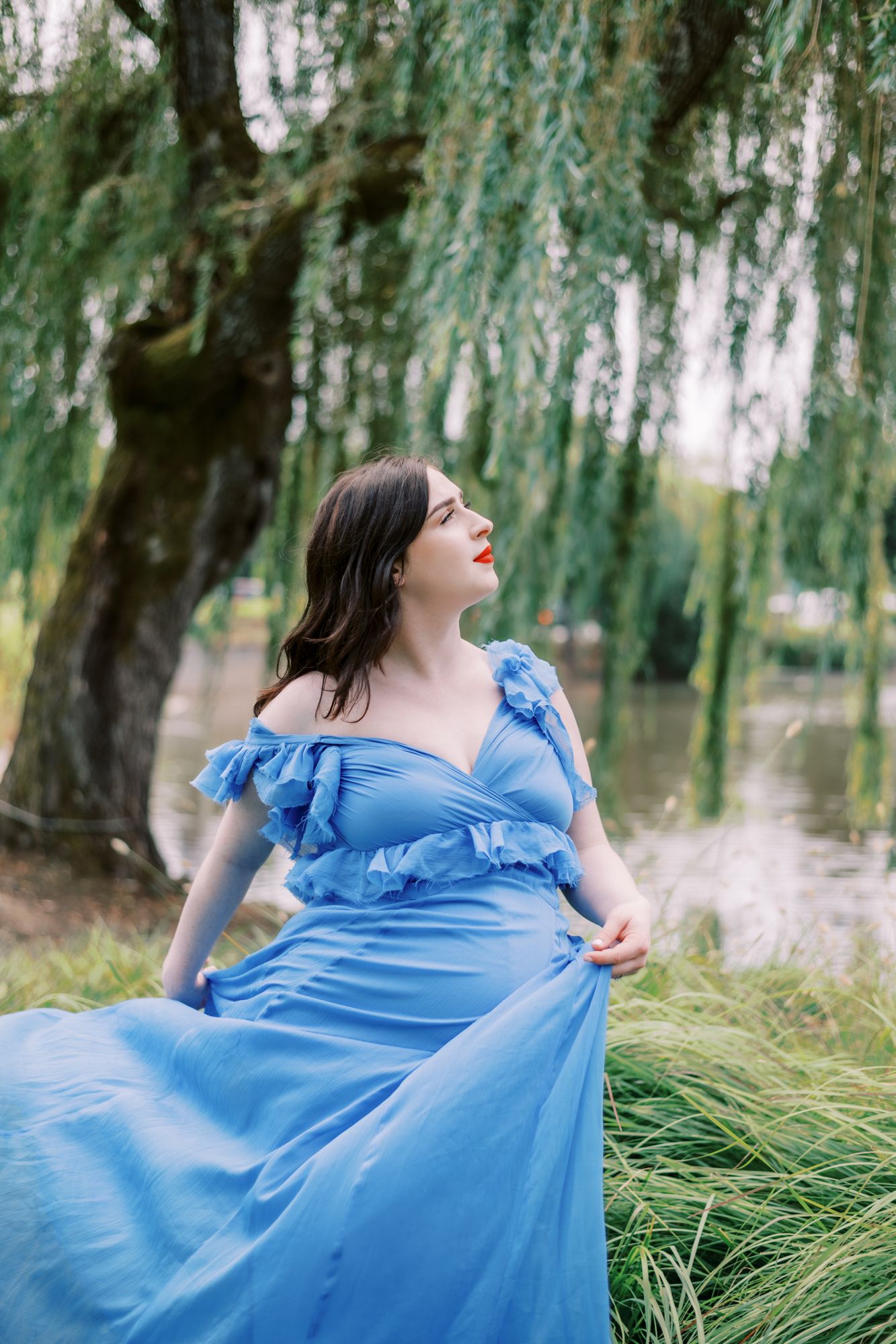 Woman in flowing blue dress poses dramatically by willow tree near water.