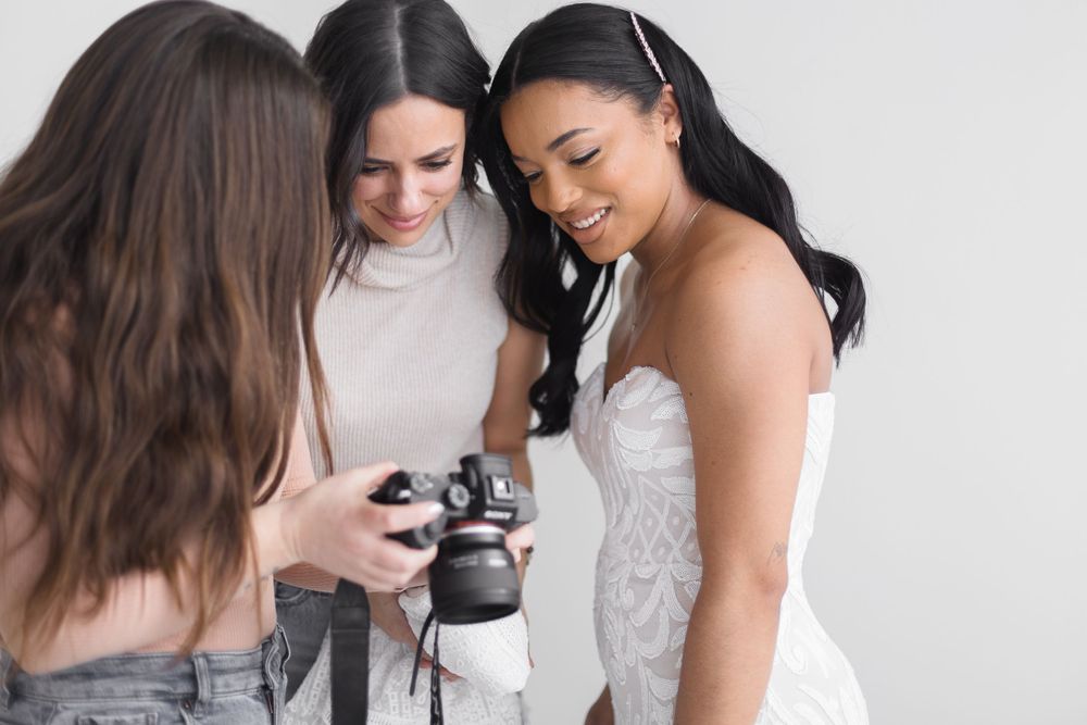 Photographer shows wedding photos on camera to smiling bride and friend in bright studio setting.