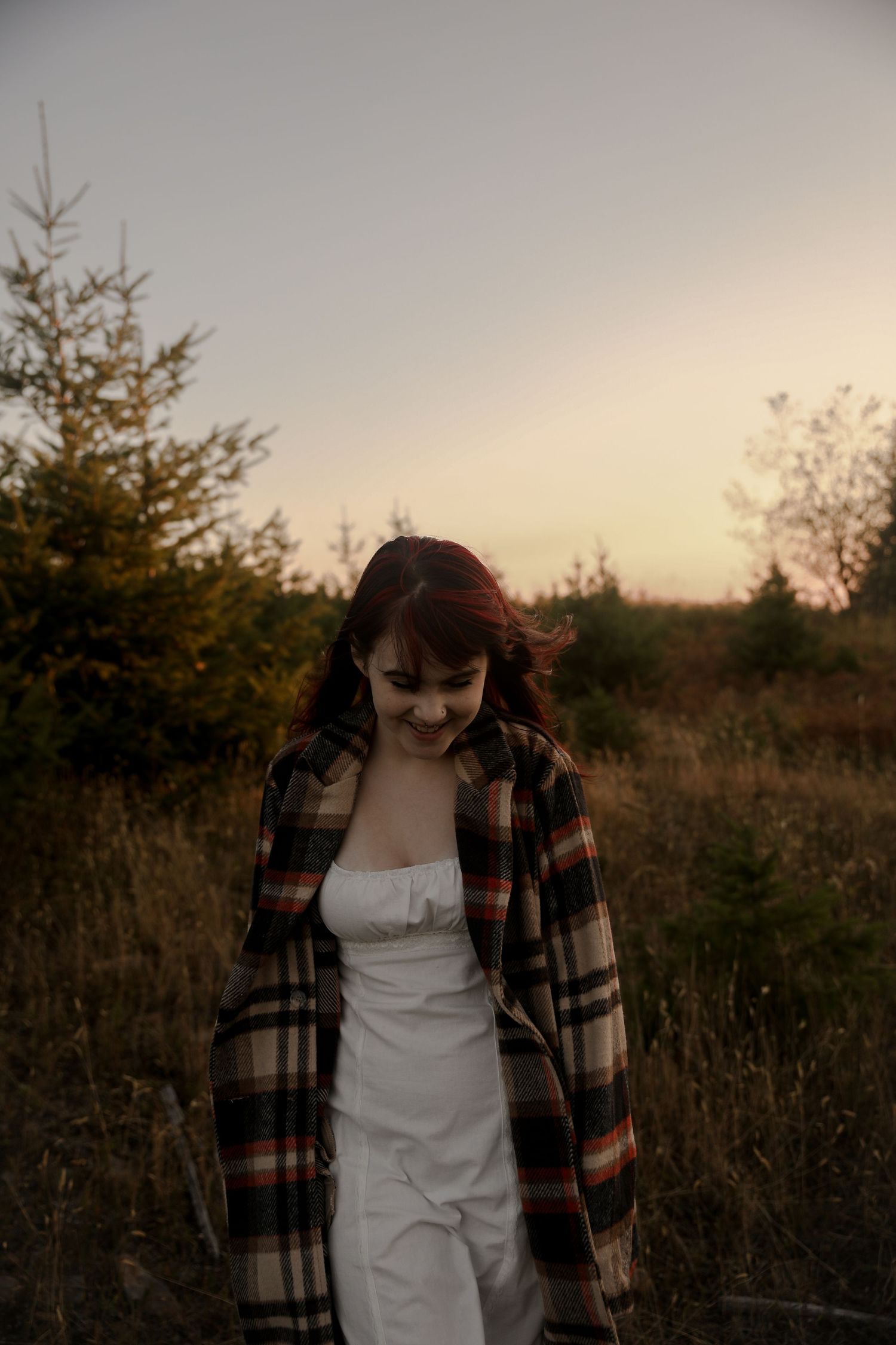 Person wearing plaid coat and white dress poses in natural setting during sunset.