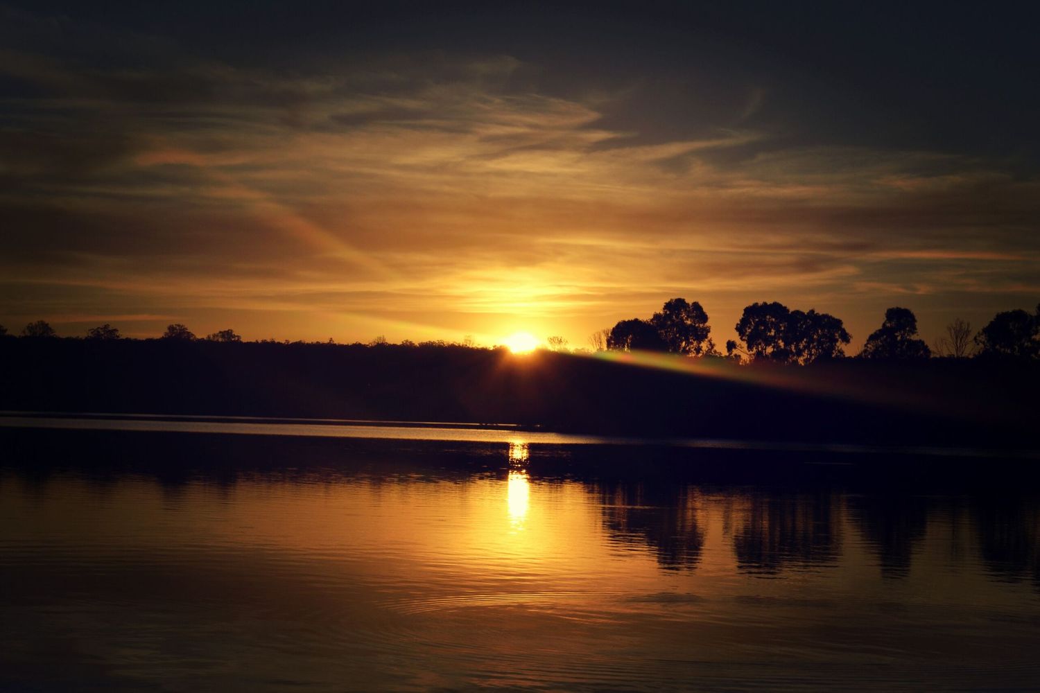 Dramatic sunset over a calm lake with silhouetted trees on the horizon.