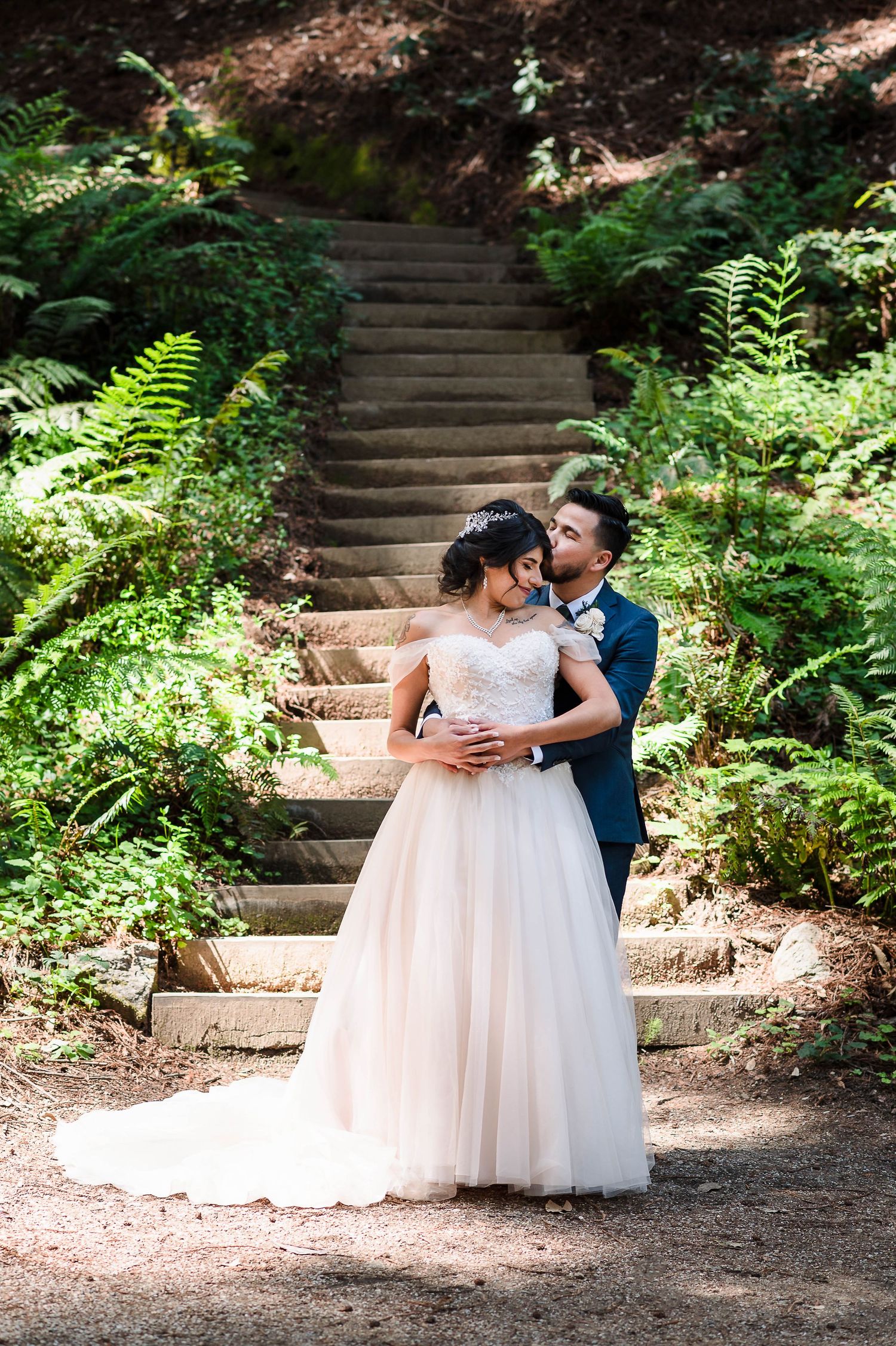 A tender embrace on rustic forest stairs surrounded by ferns and dappled sunlight.