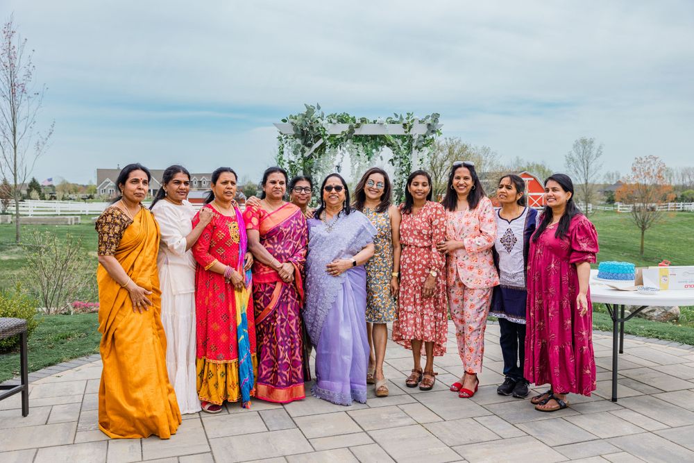 Women in vibrant saris and dresses pose together in a line under a floral arch outdoors.