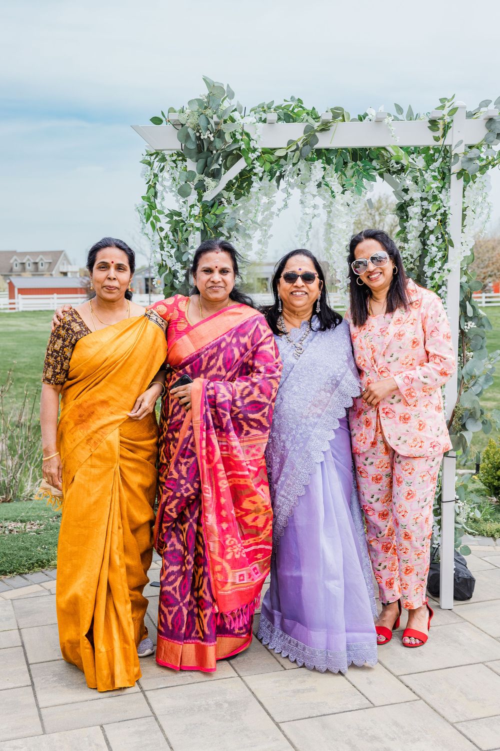 Four people in vibrant saris pose under a white arch decorated with greenery outdoors.