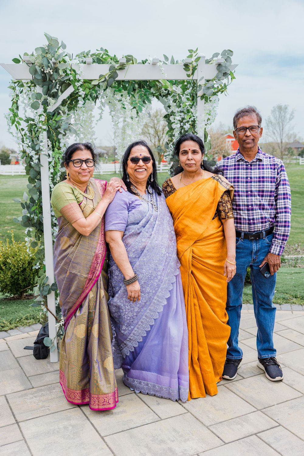 A small group in colorful Indian attire poses together under a white floral arch in a garden setting.