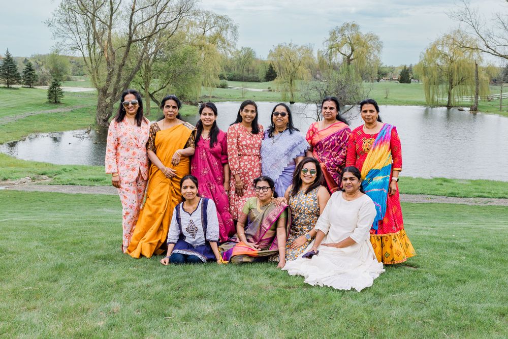 Ladies in bright traditional Indian attire pose together on grass near a pond.